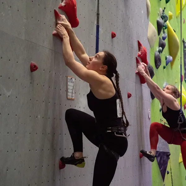 Twee vrouwen met klimgordels zijn aan het klimmen in een klimhal op een verticale wand met kleurrijke grepen. Beiden reiken omhoog, vastbesloten om de top te bereiken. Op de achtergrond is een groen deel van de klimwand te zien.
