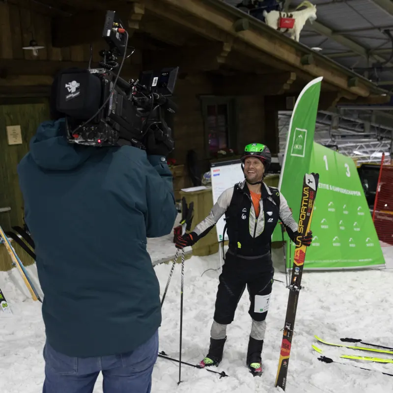 Een skiër in winterkleding glimlacht terwijl hij zijn ski’s en stokken vasthoudt; hij wordt binnenshuis op de sneeuw gefilmd door een cameraman, met op de achtergrond een groene evenementenbanner en een houten gebouw.