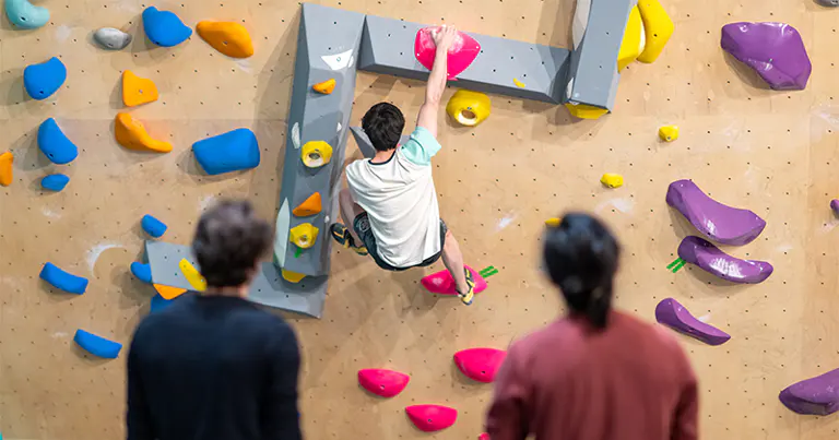 Iemand beklimt een kleurrijke bouldermuur in een zaal, terwijl twee mensen van beneden toekijken. De klimmer reikt naar een roze greep op een overhangend gedeelte.