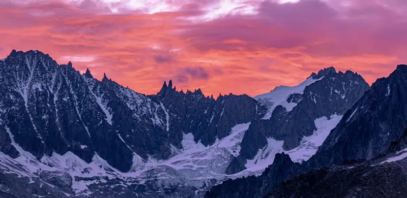 Grofgeprofileerde, met sneeuw bedekte bergen met gletsjers tegen een levendige roze en oranje zonsonderganghemel, die een dramatisch en kleurrijk alpenlandschap vormen.