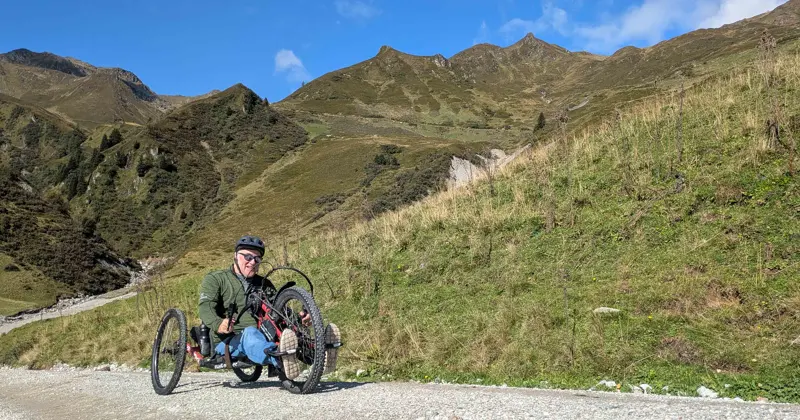 Iemand met een helm op rijdt op een handfiets over een grindpad door een bergachtig landschap met grasvelden, onder een heldere, blauwe hemel.