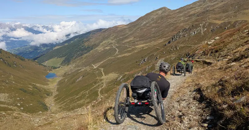 Twee mensen in terreinrolstoelen rijden een ruig bergpad op met met gras begroeide hellingen, valleien in de verte en een klein meer dat beneden zichtbaar is onder een gedeeltelijk bewolkte hemel.