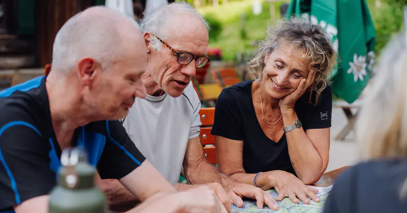Drie ouderen zitten aan een tafel buiten en bekijken samen een kaart. Eén man wijst naar de kaart, terwijl een andere man en een glimlachende vrouw vooroverbuigen en in gesprek zijn. Op de achtergrond zijn groen en parasols te zien.
