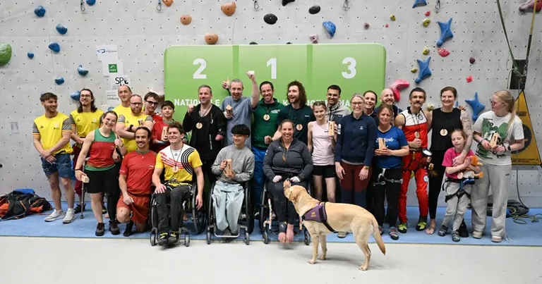 Een grote groep mensen, waaronder personen met een beperking en een blindengeleidehond, poseert en lacht samen voor een klimwand en een groen podium met de cijfers 1, 2 en 3.