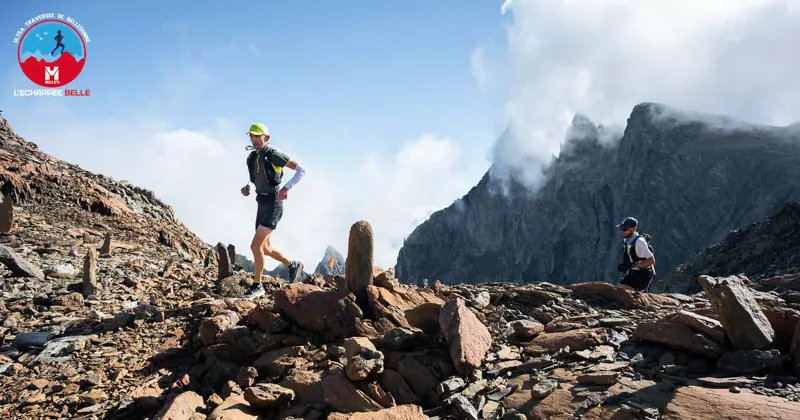 Twee trailrunners beklimmen een rotsachtig bergpad onder een heldere hemel, met indrukwekkende bergtoppen en wolken op de achtergrond. Linksboven is het logo van de race te zien.