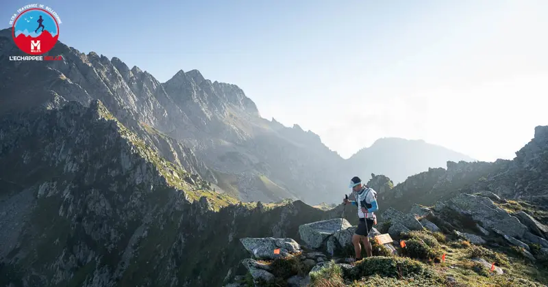 Een trailrunner staat op een rotsachtige bergkam onder een heldere hemel en kijkt uit over grillige bergtoppen en valleien. De loper draagt een rugzak en hardloopkleding; op de rotsen zijn racemarkeringen te zien. Het logo van het evenement staat in de linkerbovenhoek.