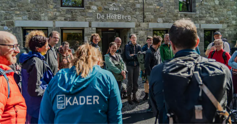 Een groep mensen met rugzakken staat op een zonnige dag glimlachend en pratend voor een stenen gebouw met het opschrift ‘De HerBerg’.