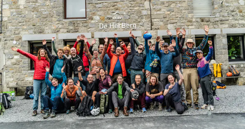 Een grote groep glimlachende mensen poseert met opgeheven armen voor een stenen gebouw met het opschrift „De HerBerg“. Velen dragen outdoor-kleding en sommigen hebben een klimhelm op.