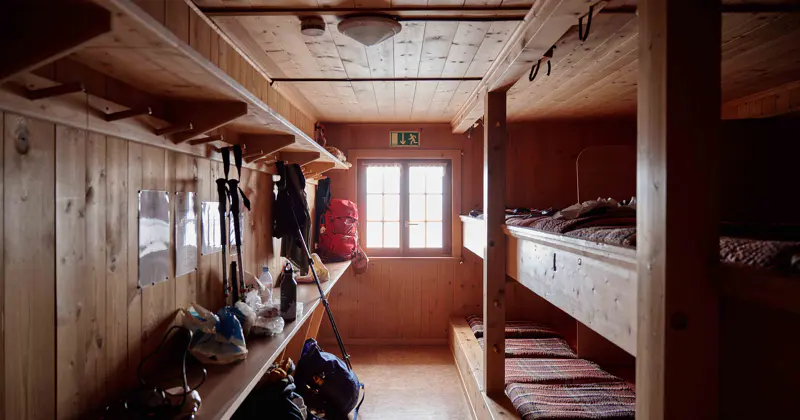 Een gezellige houten hut met stapelbedden, planken waarop uitrusting en rugzakken staan, en een bordje met ‘nooduitgang’ boven een raam dat daglicht binnenlaat.