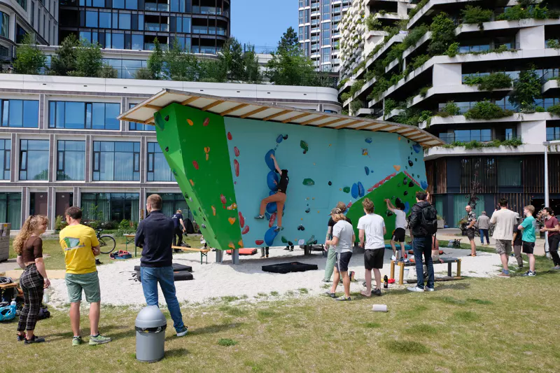 Mensen staan buiten bijeen rond een groene klimwand; één persoon is aan het klimmen terwijl de anderen toekijken. Op de achtergrond staan moderne gebouwen met veel ramen en balkons die begroeid zijn met planten.