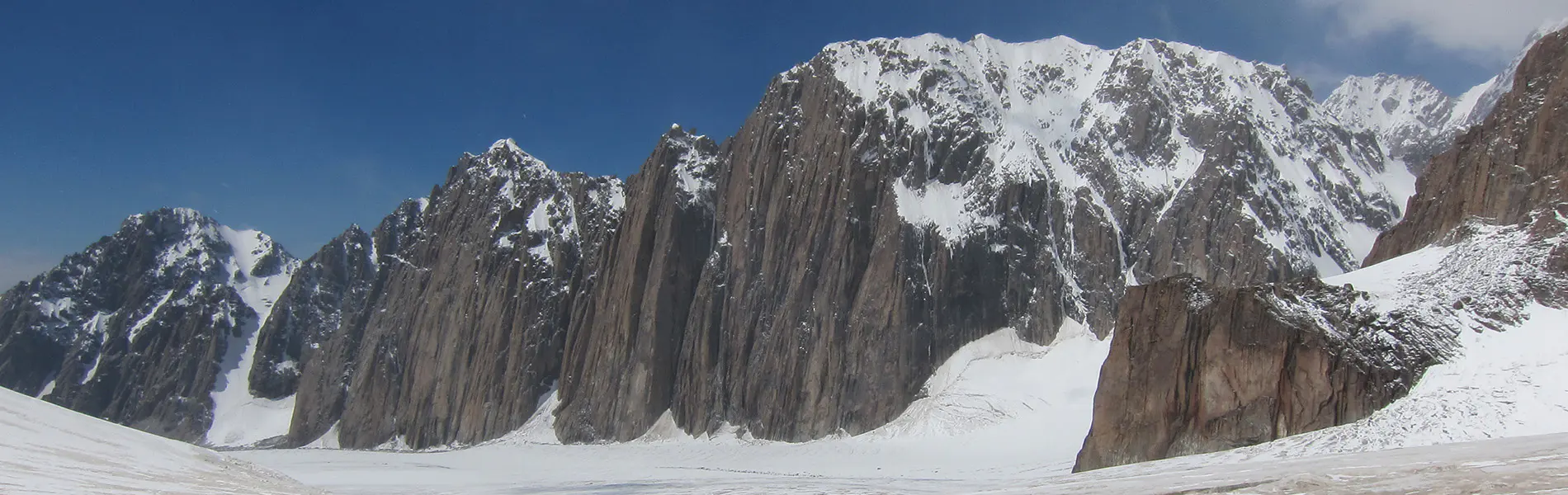 Drie bergbeklimmers in groene jassen en oranje helmen vieren feest op de top van een besneeuwde berg, terwijl ze een NKBV-vlag vasthouden, met ruige, met sneeuw bedekte bergtoppen op de achtergrond.