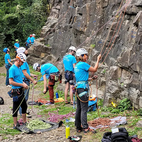 Een groep klimmers in blauwe shirts en helmen maakt touwen en uitrusting klaar aan de voet van een rotsachtige klimwand in de open lucht, omgeven door groen.